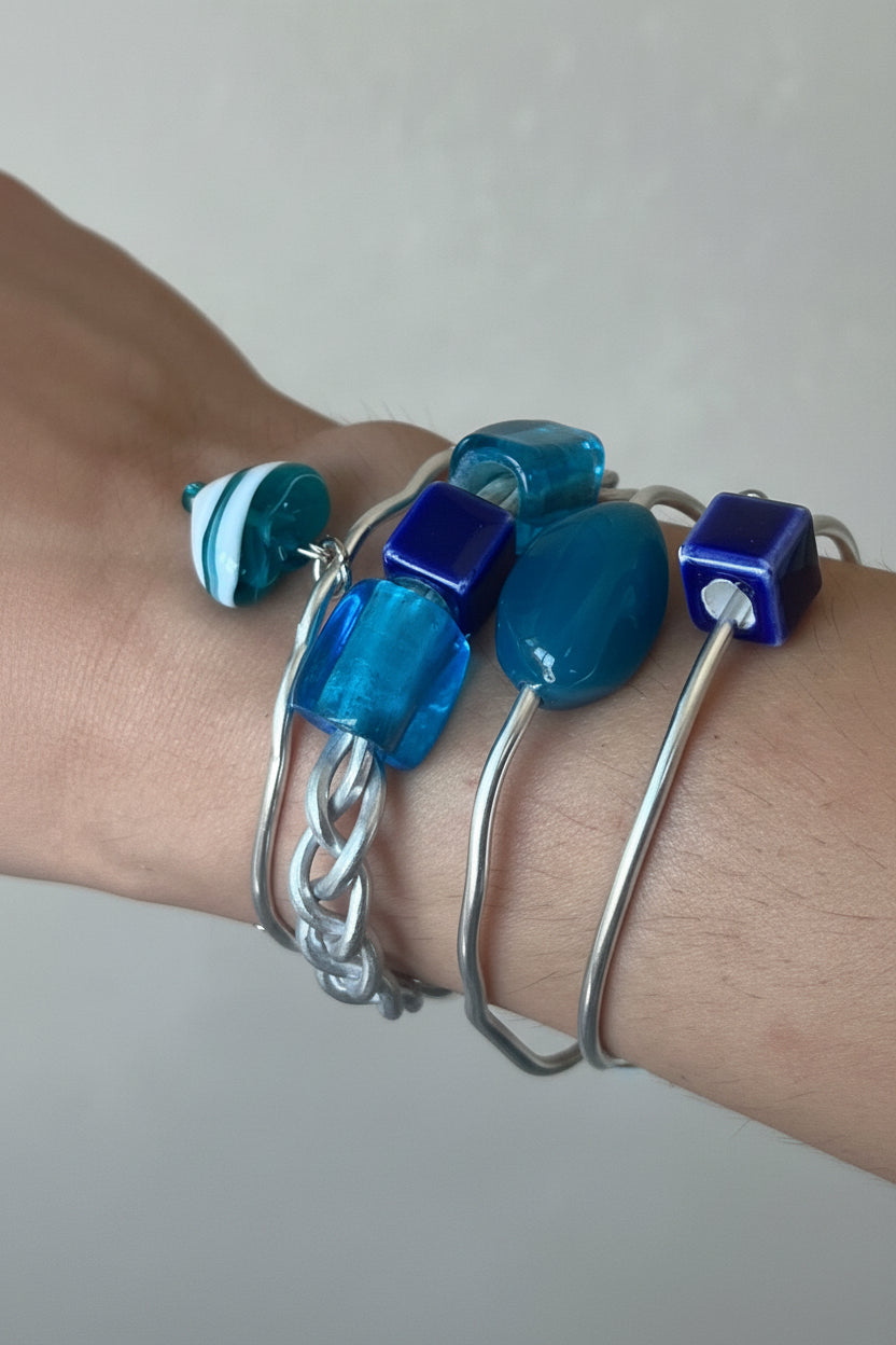 A close-up of a person's wrist wearing a stack of handmade blue and silver bracelets, including glass beads, a chain link, and a striped charm, against a blurred tiled background.