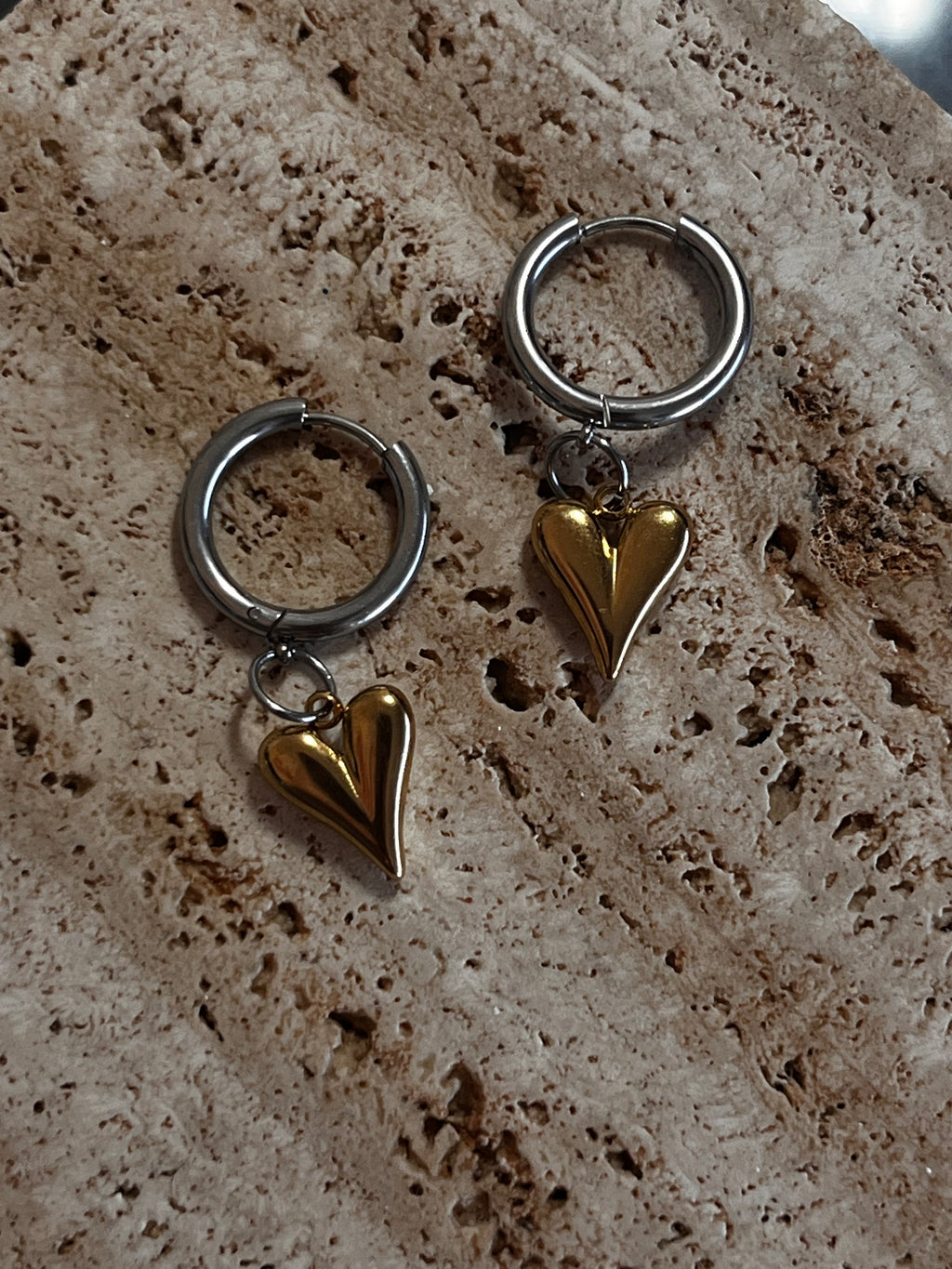 Two silver hoop earrings with gold heart charms on a textured stone surface.