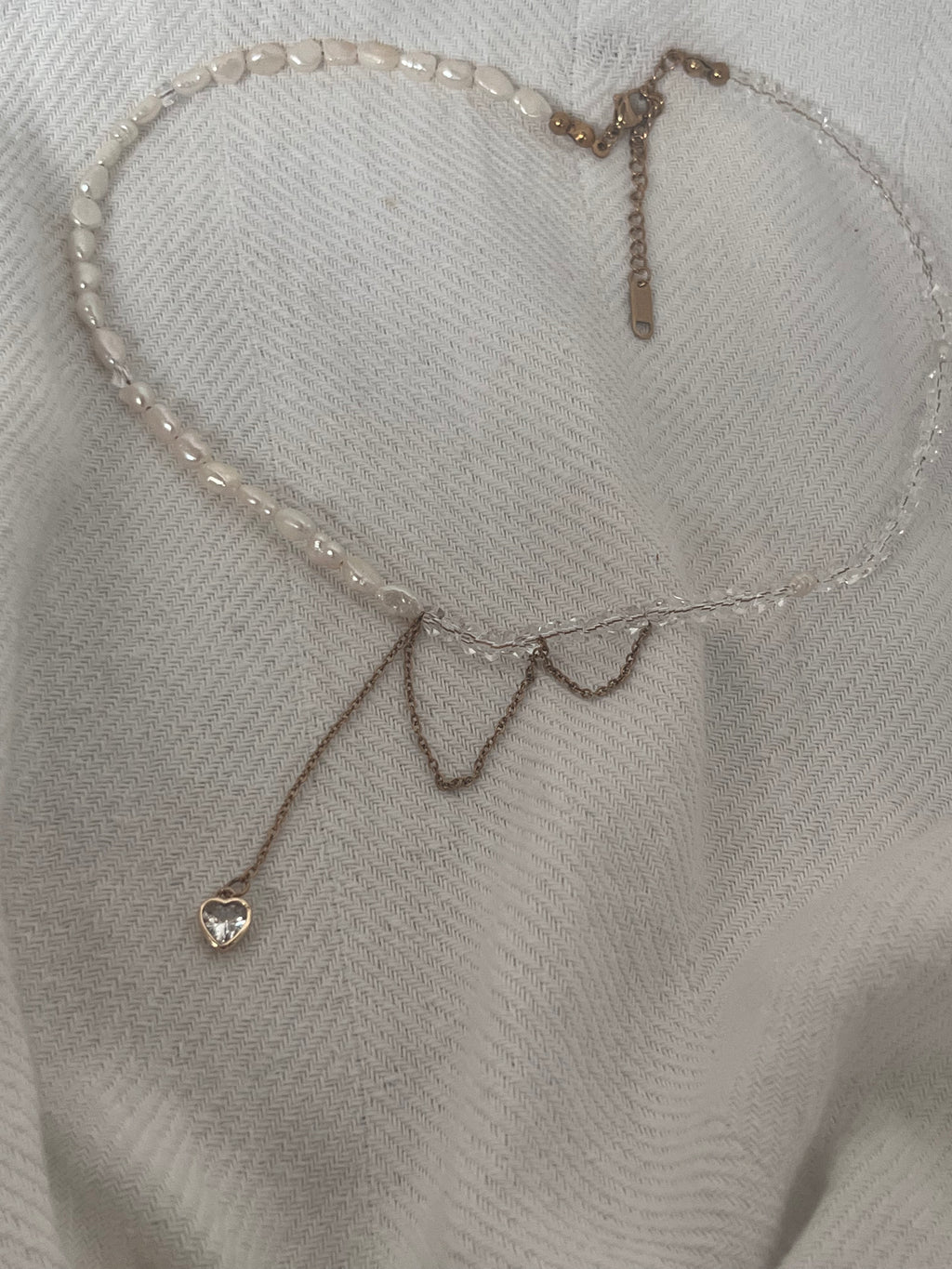 Handmade asymmetric necklace featuring white pearls, faceted crystals, and a crystal heart pendant on a soft white fabric background.