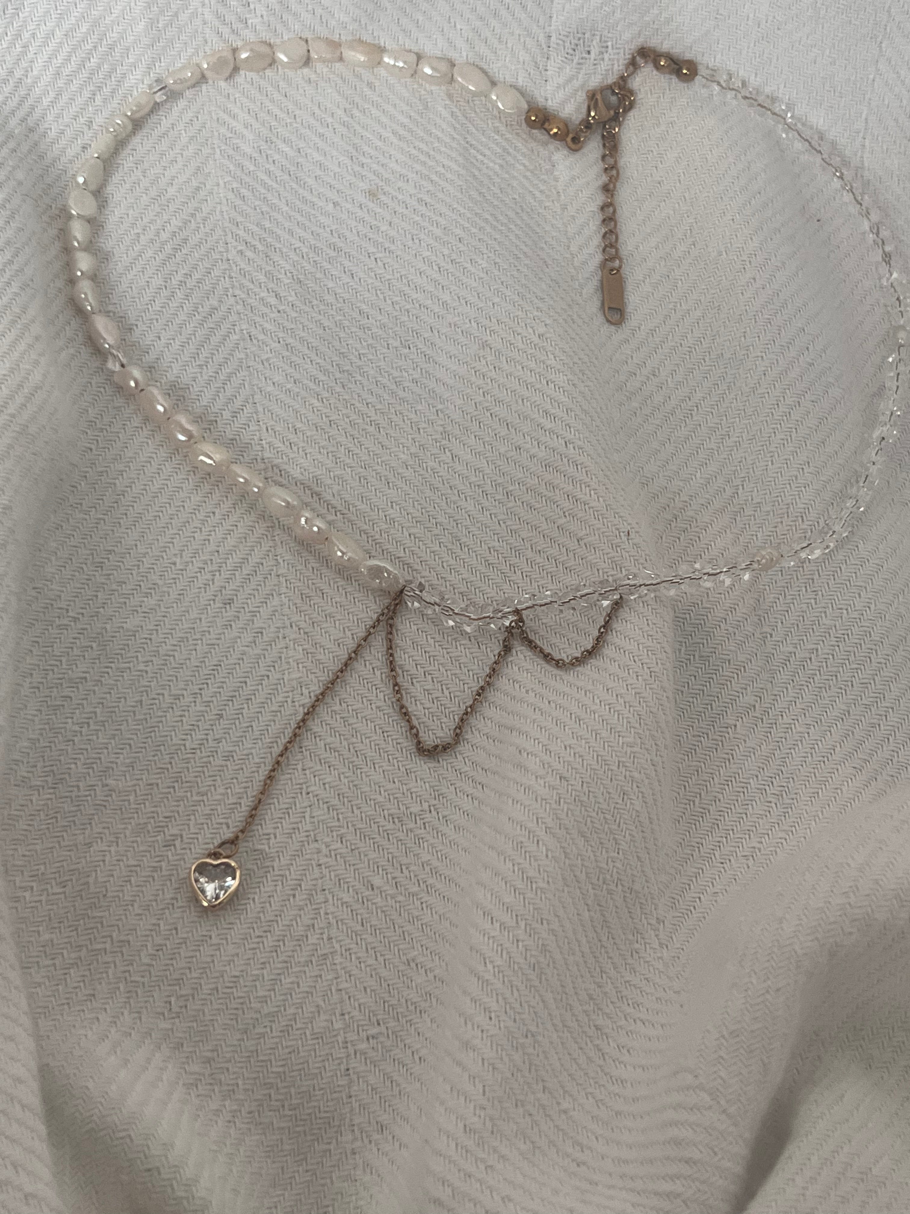 Handmade asymmetric necklace featuring white pearls, faceted crystals, and a crystal heart pendant on a soft white fabric background.